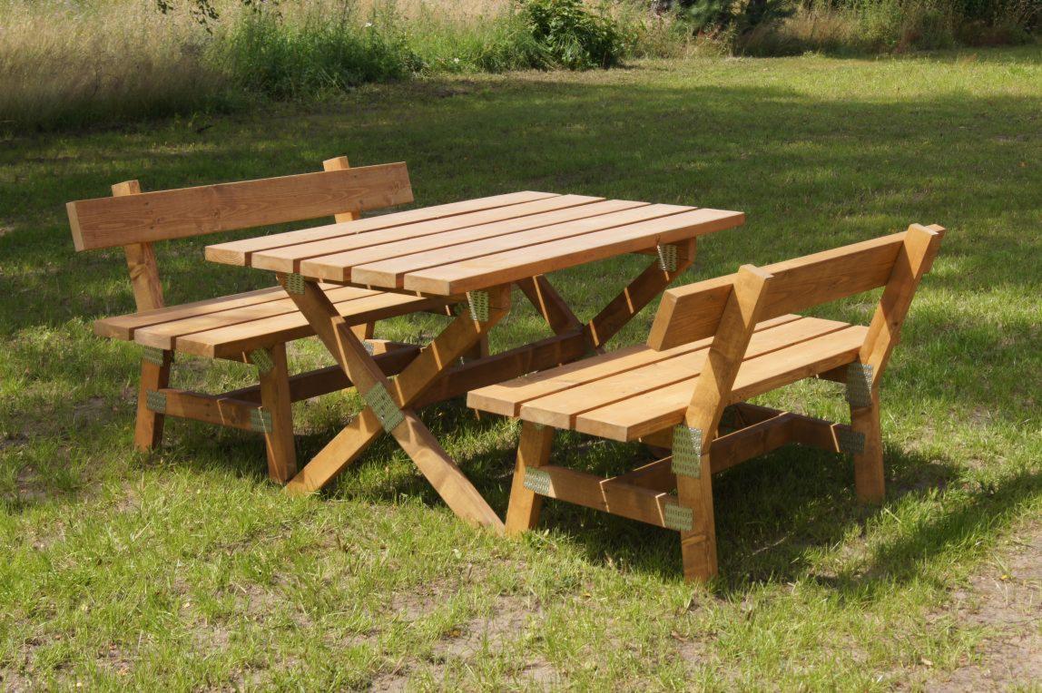 Drewniany zestaw mebli ogrodowych: stół z ławkami, wykonany z jasnego drewna, z widocznymi metalowymi wzmocnieniami, ustawiony na trawniku w słoneczny dzień.