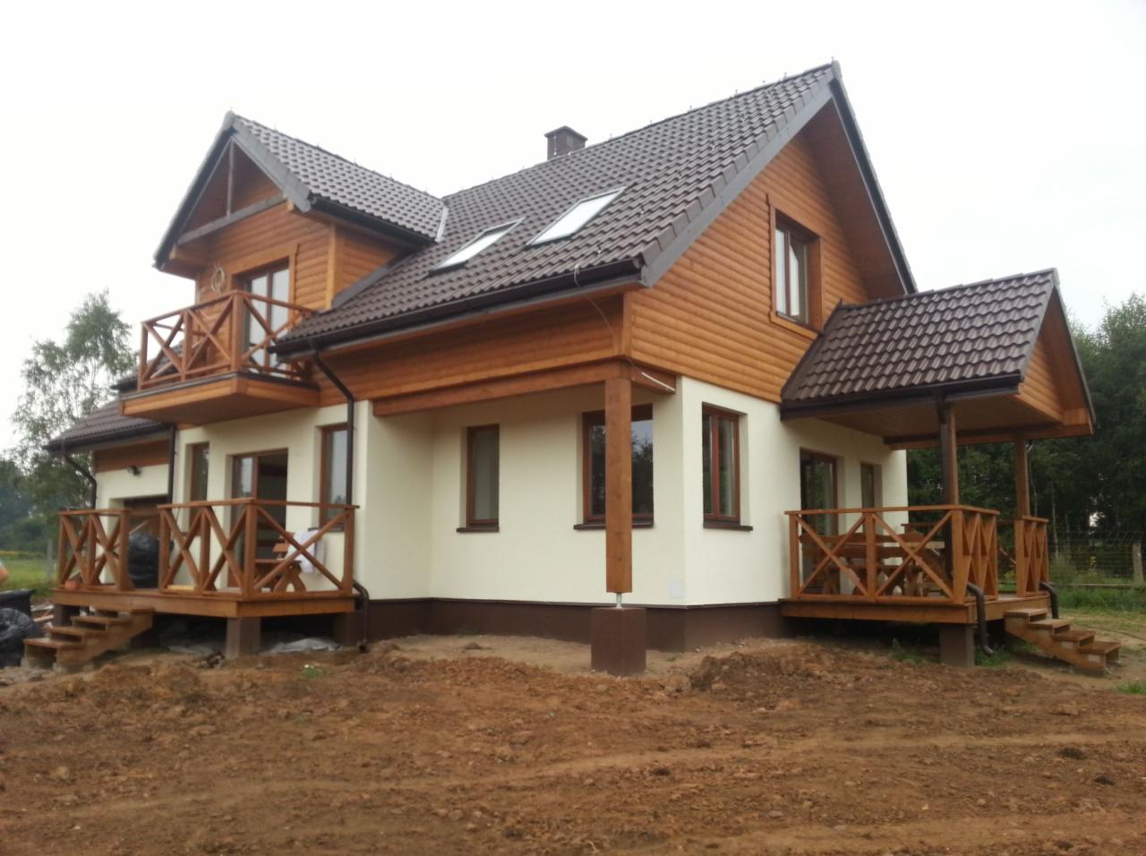 Dwukondygnacyjny dom drewniany z brązowym dachem, balkonem i werandą, otoczony ziemią w trakcie prac wykończeniowych.