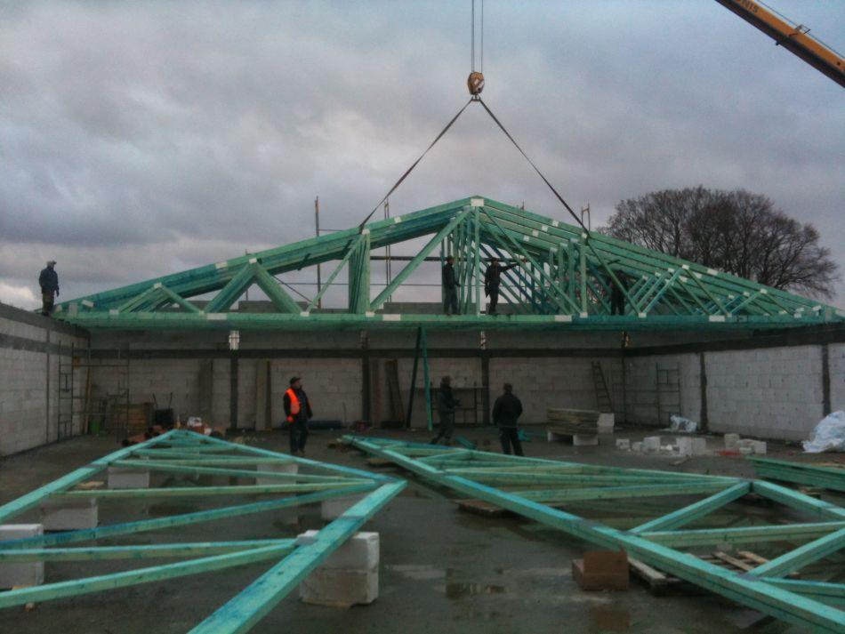 Montaż prefabrykowanych wiązarów dachowych z drewna o zielonkawym odcieniu na budowie hali; widoczny dźwig w tle i pracownicy w trakcie montażu.