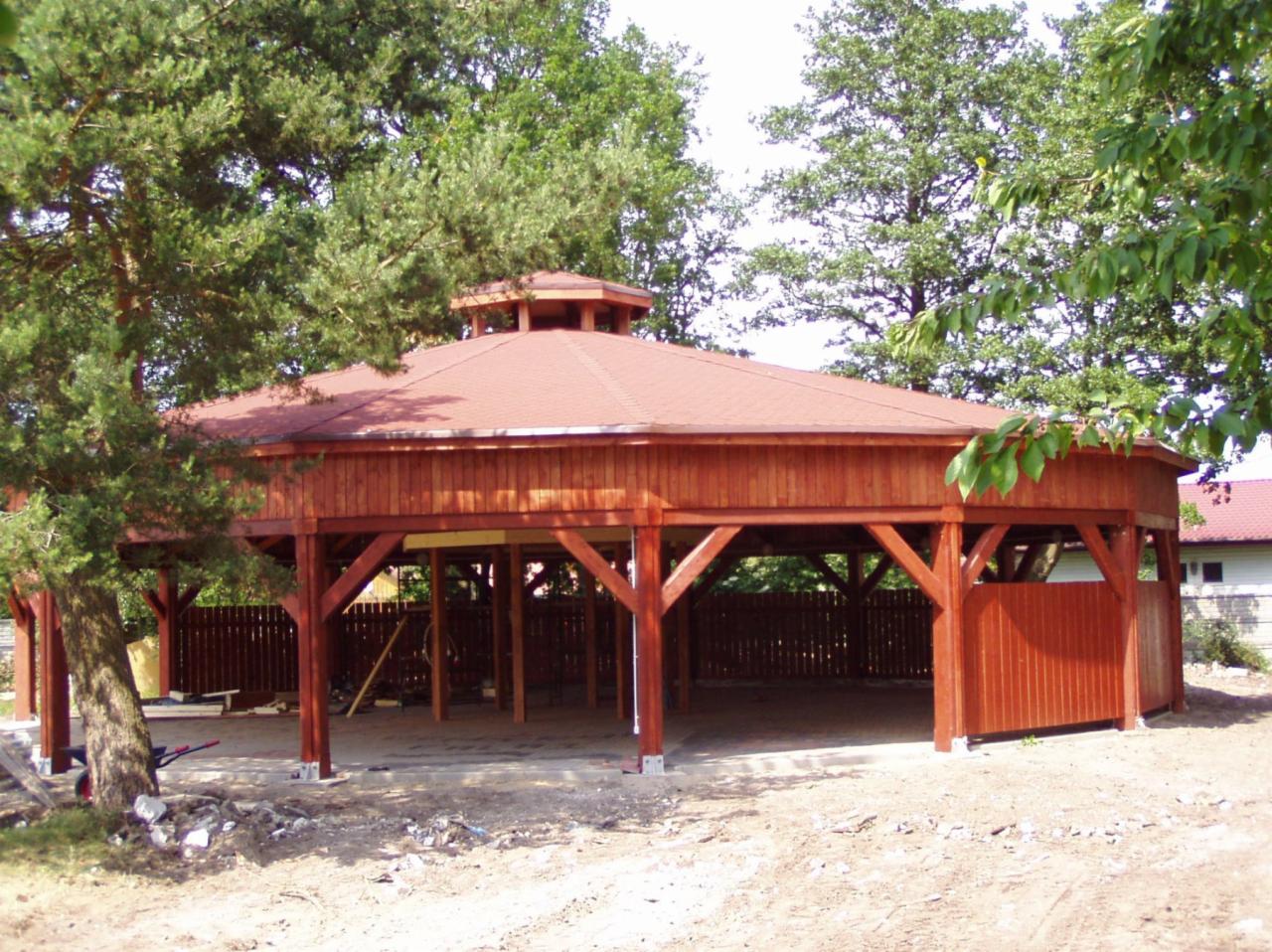Ośmiokątna, drewniana altana z czerwonym dachem, częściowo osłonięta drewnianą ścianką, na betonowej wylewce, otoczona zielenią.