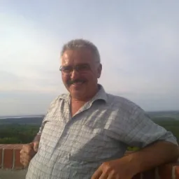 Uśmiechnięty mężczyzna w okularach opiera się o ceglany murek z widokiem na rozległy krajobraz z lasem i jeziorem w tle. Słoneczny dzień.