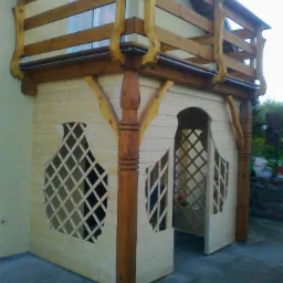 Drewniana konstrukcja balkonu z ażurowymi ścianami, wykonana z jasnego drewna, z ciemniejszymi, toczonymi kolumnami i balustradą, umieszczona przy jasnej ścianie budynku.