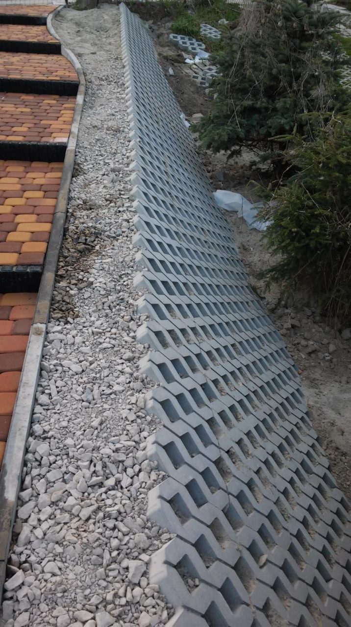 Ułożona szara ażurowa kostka betonowa na zboczu, obok schodów z czerwono-brązowej kostki. Widoczne kamienie i roślinność. Kątowe ujęcie.