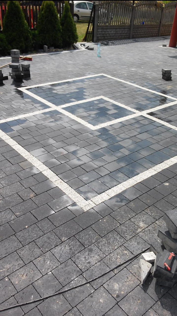 Ułożona, grafitowa kostka brukowa z białymi, kwadratowymi obramowaniami. Częściowo mokra nawierzchnia odbija niebo. W tle zieleń i ogrodzenie.