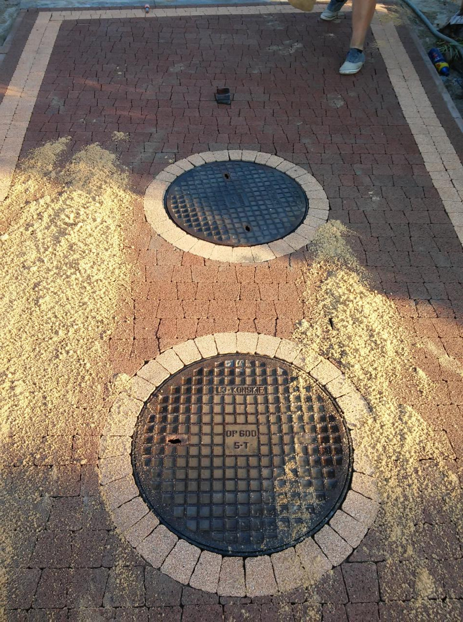 Ułożona kostka brukowa w odcieniach brązu z dwoma studzienkami kanalizacyjnymi, wykończonymi okręgami z jaśniejszej kostki. Widoczny piasek na powierzchni.