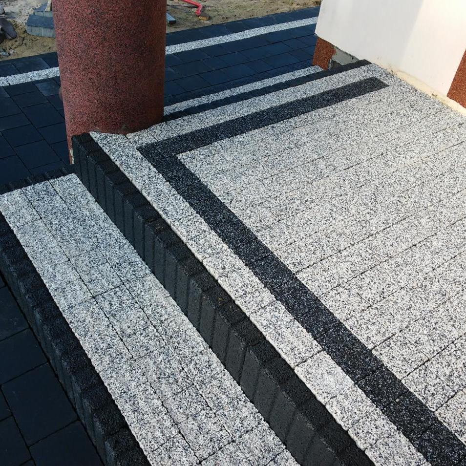 Schody z kostki granitowej w odcieniach szarości i czerni, z kolumną z czerwonego granitu po lewej stronie. Detal wykończenia z bliska, widoczne fugi i faktura kamienia.