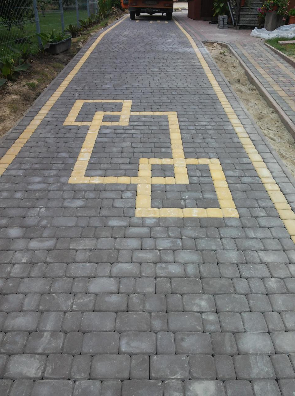 Ułożona szara kostka brukowa z żółtym wzorem geometrycznym na podjeździe. Widoczny fragment chodnika z kostki i zieleń. W tle zaparkowany samochód.