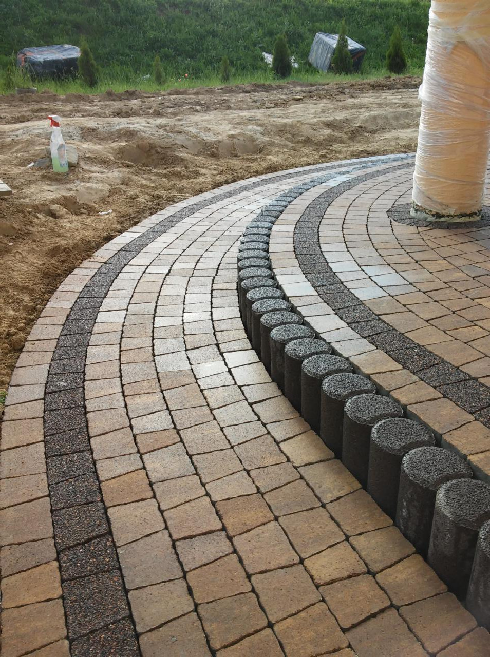 Ułożona kostka brukowa w łukowaty wzór z obrzeżami z kostki granitowej. Częściowo widoczny trawnik i kolumna owinięta folią. Prace brukarskie w toku.