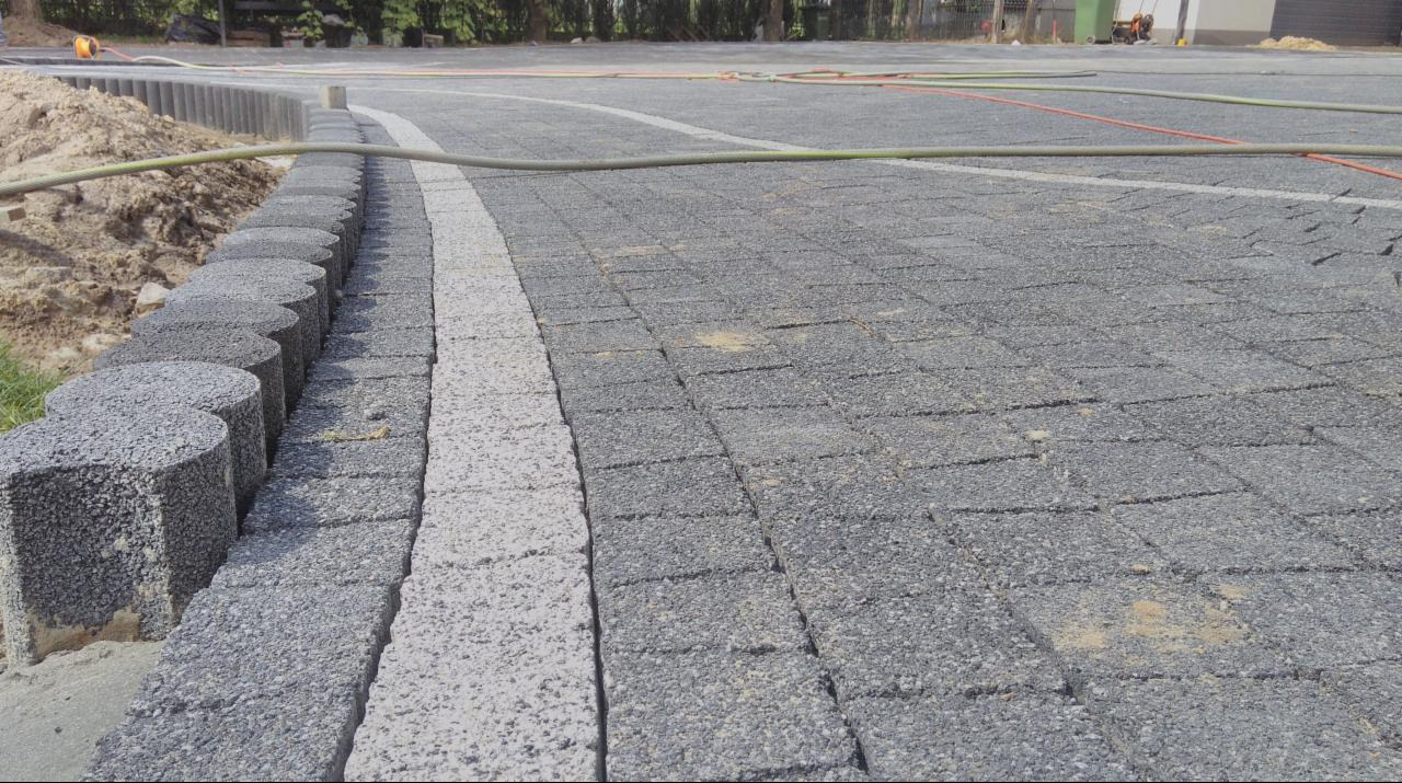 Ułożona szara kostka brukowa z jasnym pasem i obrzeżem z okrągłych elementów. Widoczne przygotowane podłoże i sprzęt budowlany w tle. Kąt ujęcia z dołu podkreśla perspektywę.