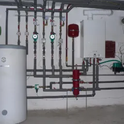 Kotłownia z białym bojlerem marki Biwar, piecem gazowym De Dietrich, licznymi rurami, zaworami, pompami obiegowymi i zbiornikami wyrównawczymi w kolorach zielonym i czerwonym, zamontowana na białej...