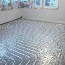 Białe rury ogrzewania podłogowego ułożone na siatce w surowym pomieszczeniu, widoczne okna i materiały budowlane w tle.