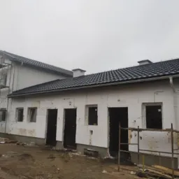 Budynek w trakcie ocieplania, widoczne białe płyty styropianowe na ścianach, nowy, ciemny dach z kominami, rusztowania przy ścianie szczytowej, pochmurne niebo.