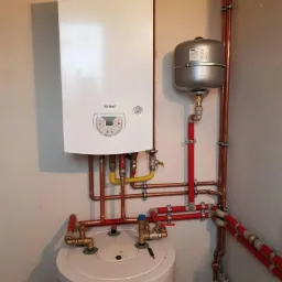 Biały kondensacyjny piec gazowy Termet z podłączeniami z miedzi i czerwonych rur PEX, zawór bezpieczeństwa i szary zbiornik wyrównawczy. Montaż w rogu pomieszczenia.