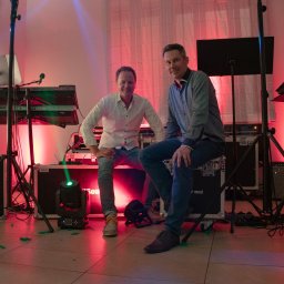 AdiSound Adrian Górczyński - Dwóch mężczyzn siedzi na skrzyniach sprzętu muzycznego w Skórzewie, z widocznymi instrumentami klawiszowymi, oświetleniem scenicznym i wzmacniaczami, w tle zasłona i ceglany fragment ściany.