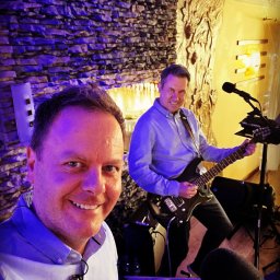 AdiSound Adrian Górczyński - Selfie z gitarzystą w tle podczas występu; widoczny mikser, mikrofon i dekoracyjna ściana z kamienia podświetlona na niebiesko.