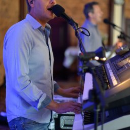 AdiSound Adrian Górczyński - Mężczyzna w niebieskiej koszuli śpiewa do mikrofonu i gra na keyboardzie podczas występu, w tle drugi muzyk.