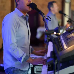 Mężczyzna w niebieskiej koszuli śpiewa do mikrofonu i gra na keyboardzie podczas występu, w tle drugi muzyk.