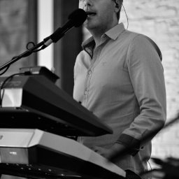 AdiSound Adrian Górczyński - Czarno-białe zdjęcie muzyka ze Skórzewa śpiewającego do mikrofonu i grającego na keyboardzie. Widoczny fragment ceglanej ściany w tle.