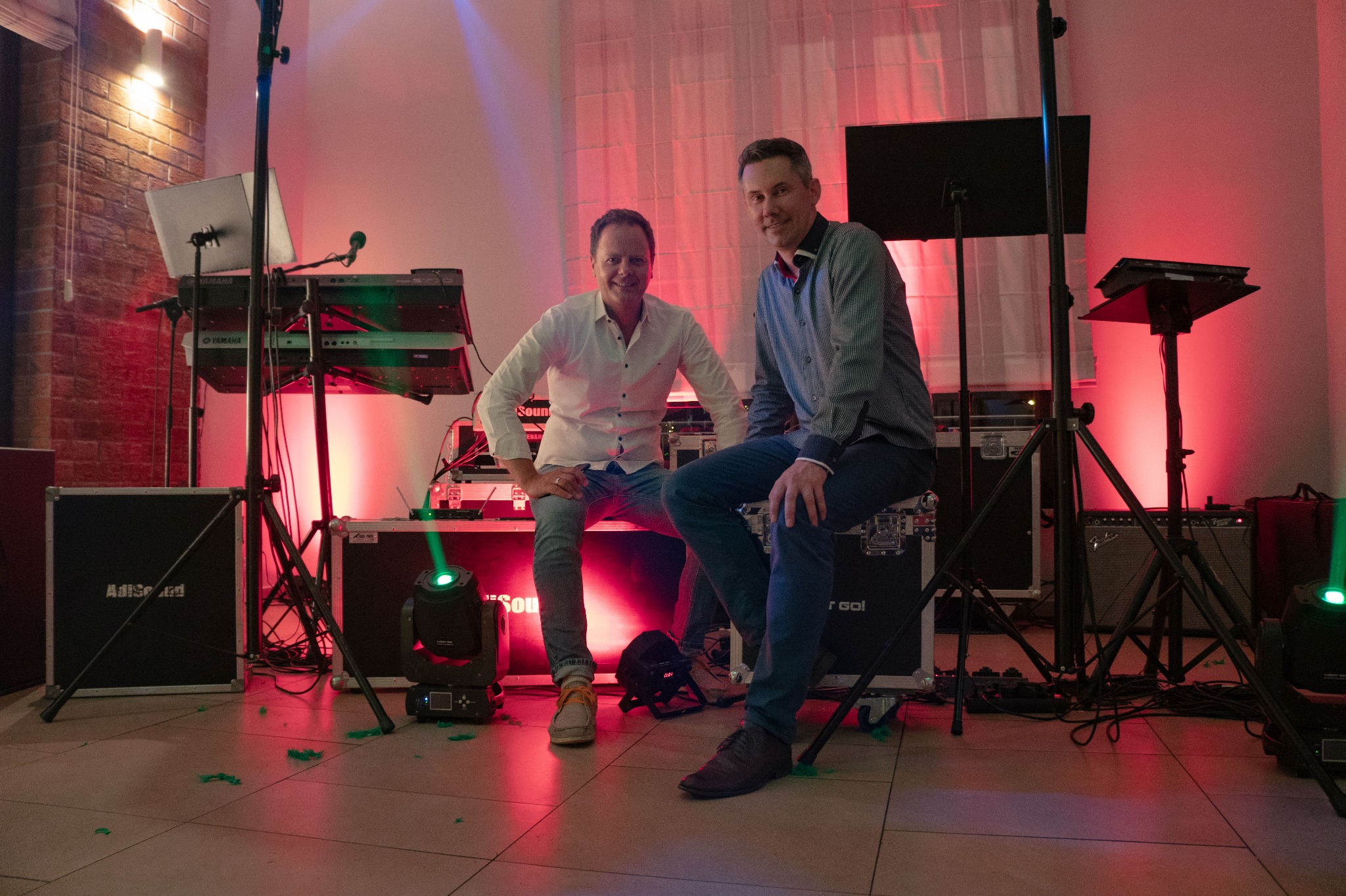 Dwóch mężczyzn siedzi na skrzyniach sprzętu muzycznego w Skórzewie, z widocznymi instrumentami klawiszowymi, oświetleniem scenicznym i wzmacniaczami, w tle zasłona i ceglany fragment ściany.