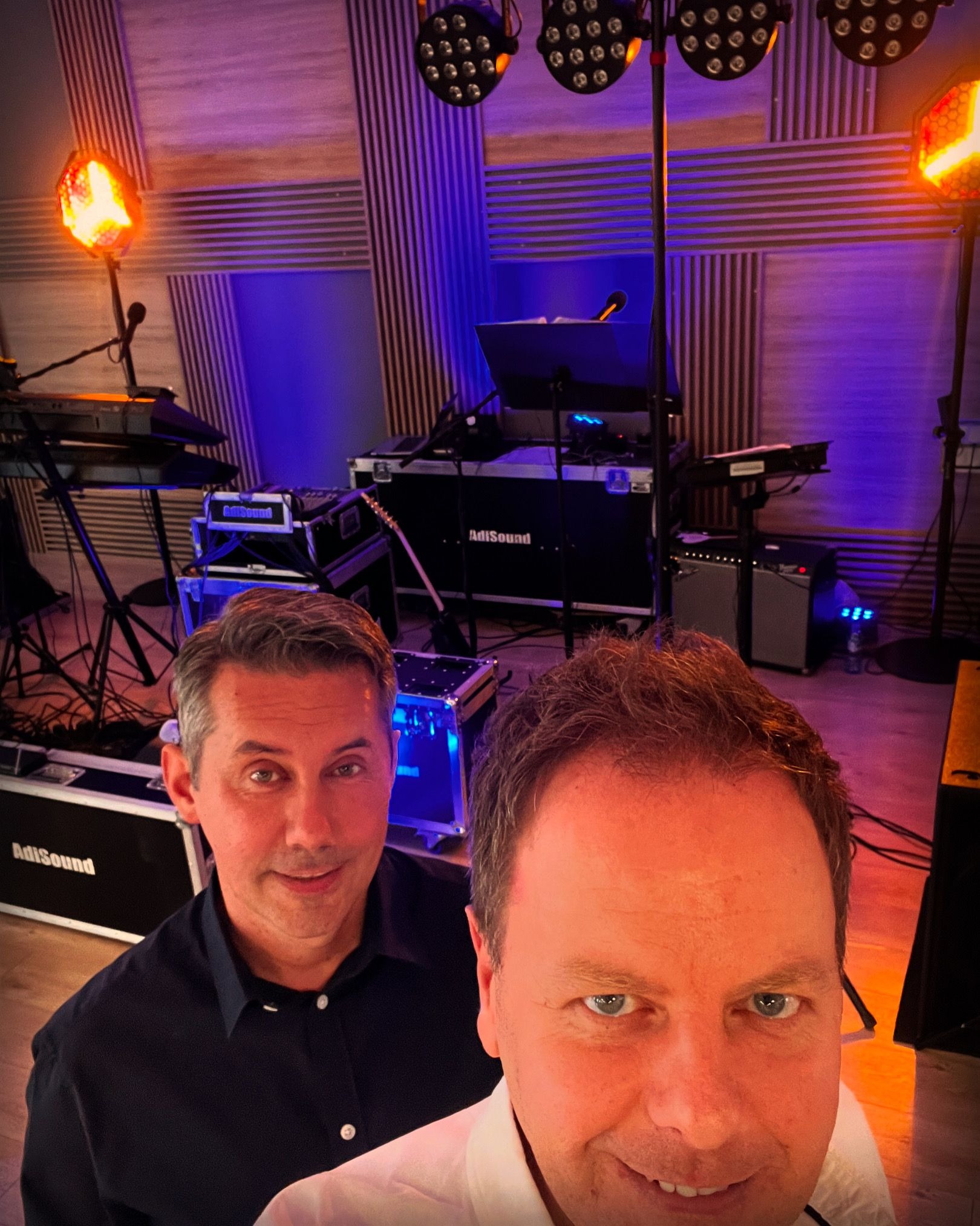 Selfie dwóch mężczyzn na tle sceny z instrumentami, oświetleniem estradowym i sprzętem nagłośnieniowym marki AdiSound.