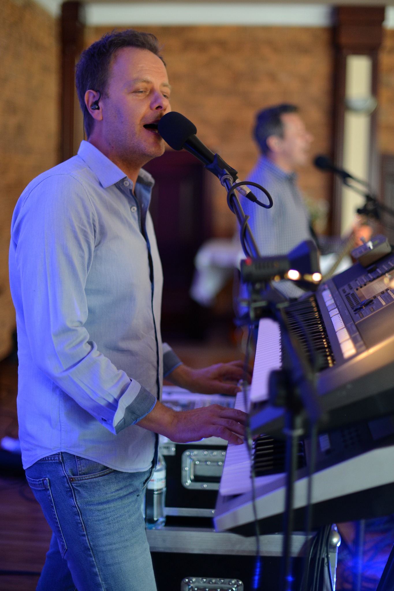 Mężczyzna w niebieskiej koszuli śpiewa do mikrofonu i gra na keyboardzie podczas występu, w tle drugi muzyk.