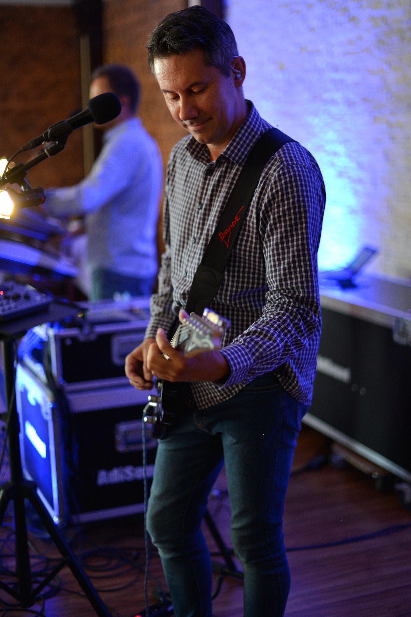 Gitarzysta z gitarą elektryczną Ibanez grający na scenie w koszuli w kratę i dżinsach, w tle mikrofon i klawiszowiec w niebieskim oświetleniu.