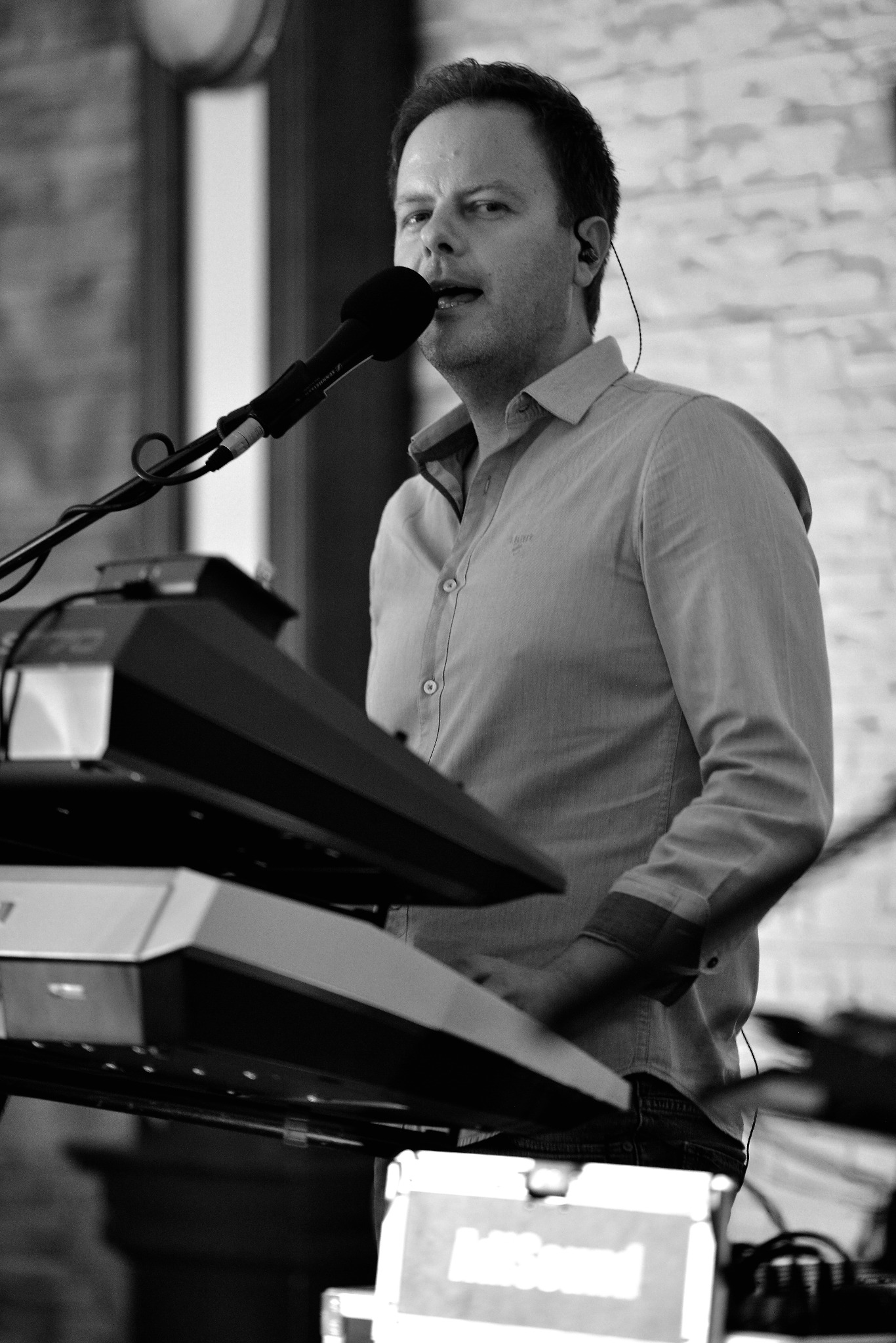 Czarno-białe zdjęcie muzyka ze Skórzewa śpiewającego do mikrofonu i grającego na keyboardzie. Widoczny fragment ceglanej ściany w tle.