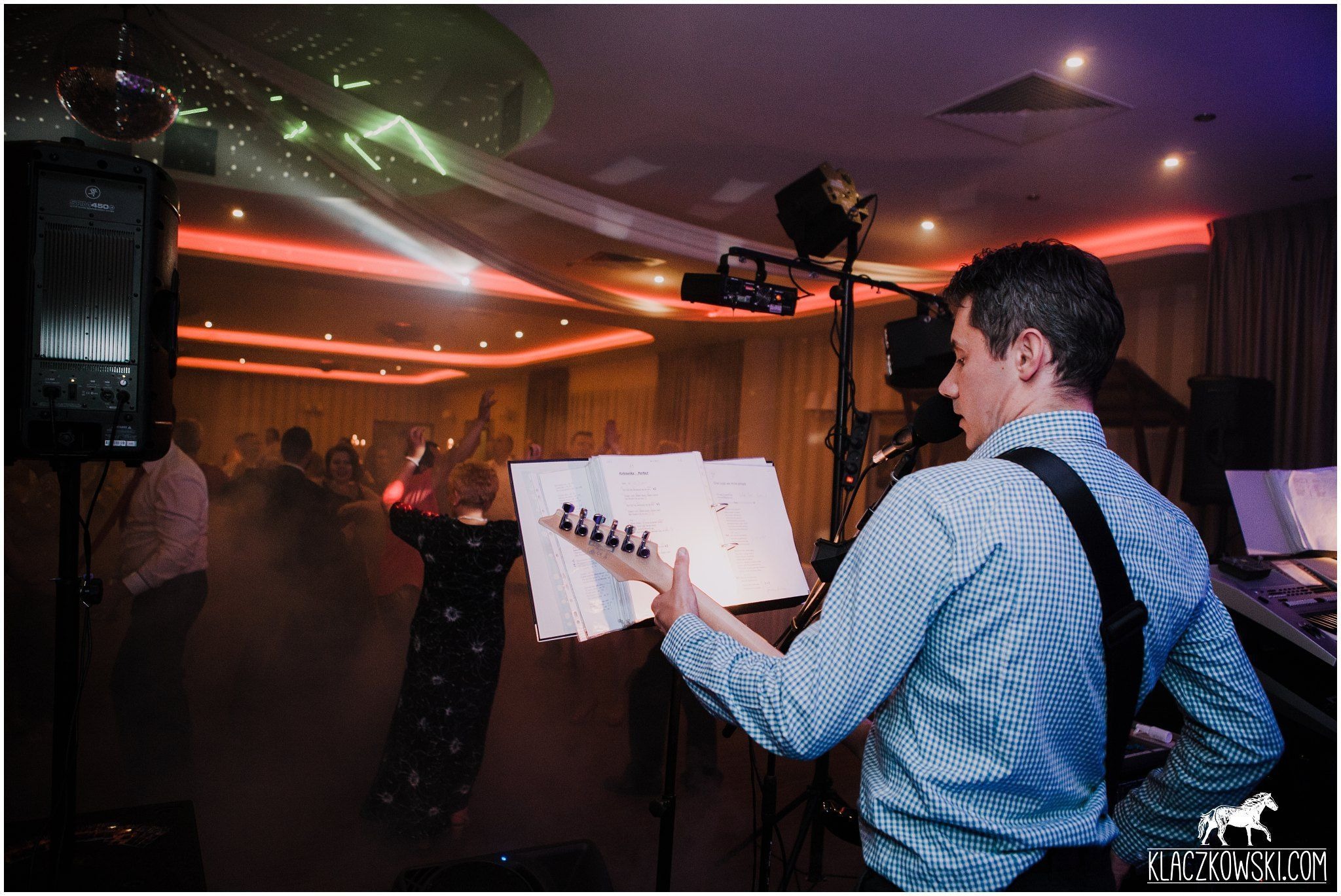 Gitarzysta w niebieskiej koszuli grający na gitarze elektrycznej podczas występu na tle tańczących osób i oświetlenia scenicznego, nuty na statywie.