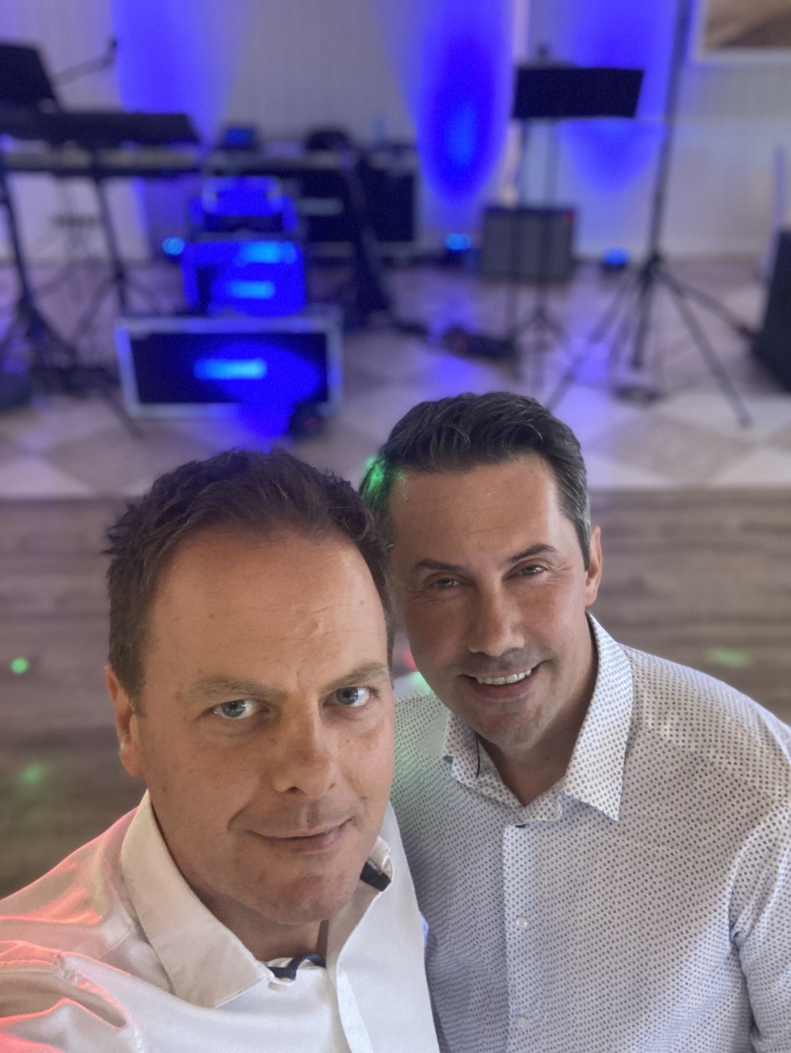 Selfie dwóch uśmiechniętych mężczyzn na tle rozmytej sceny z instrumentami i oświetleniem scenicznym w odcieniach niebieskiego.