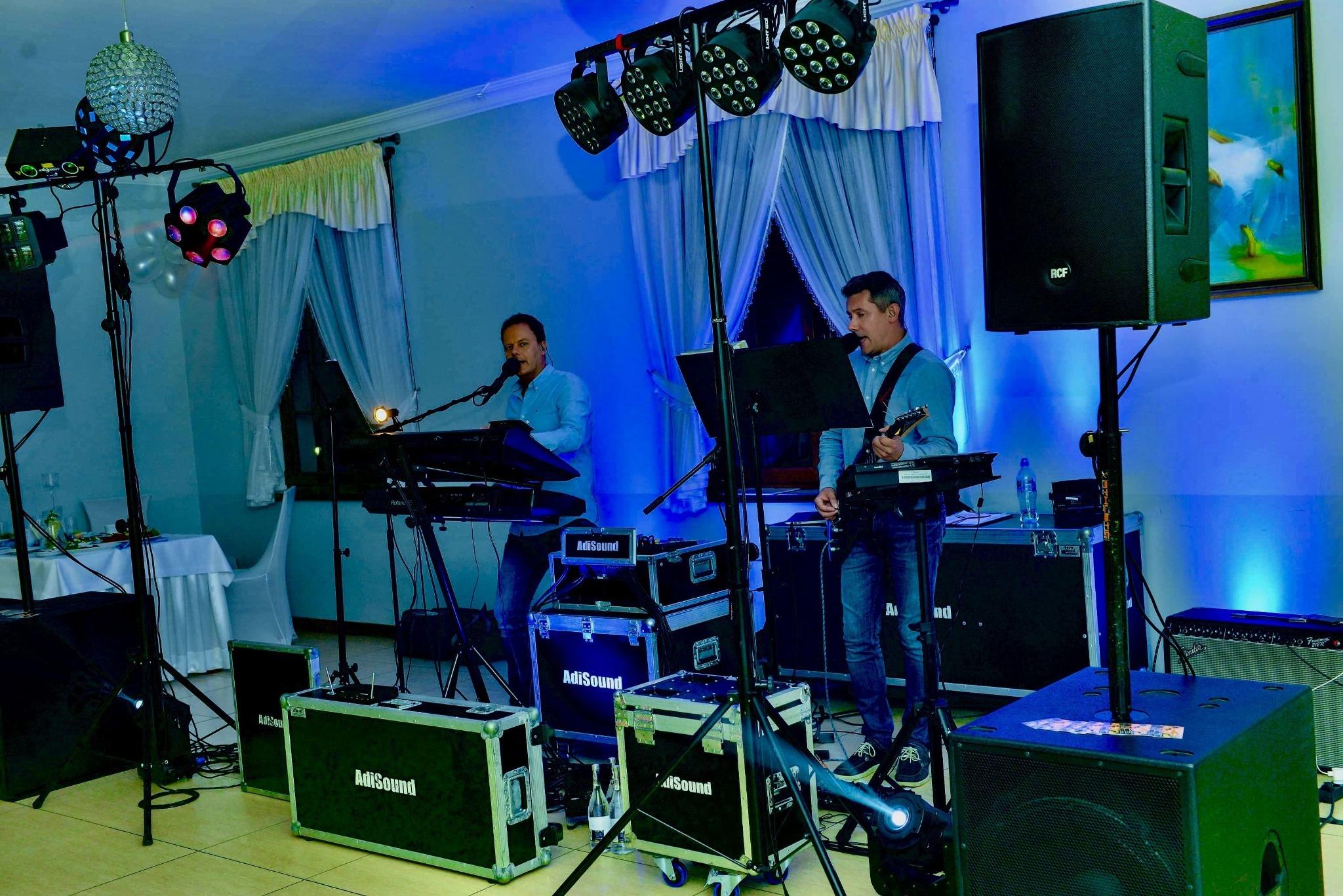 Dwóch muzyków grających na instrumentach (keyboard i gitara) podczas występu na tle sali z dekoracjami i oświetleniem scenicznym, widoczne skrzynie z logo AdiSound.