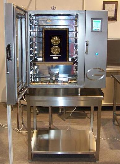 Profesjonalna, metalowa komora wędzarnicza z otwartymi drzwiami, prezentująca wewnątrz oprawione medale, z panelem sterowania i logiem firmy na boku, umieszczona na metalowym stole.