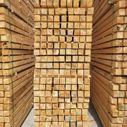 Stosy świeżo przetartego, jasnego drewna sosnowego w postaci krawędziaków 100x100, ułożone w regularne pryzmy, widoczne ślady cięcia i naturalny rysunek drewna, niektóre krawędziaki z kolorowymi...