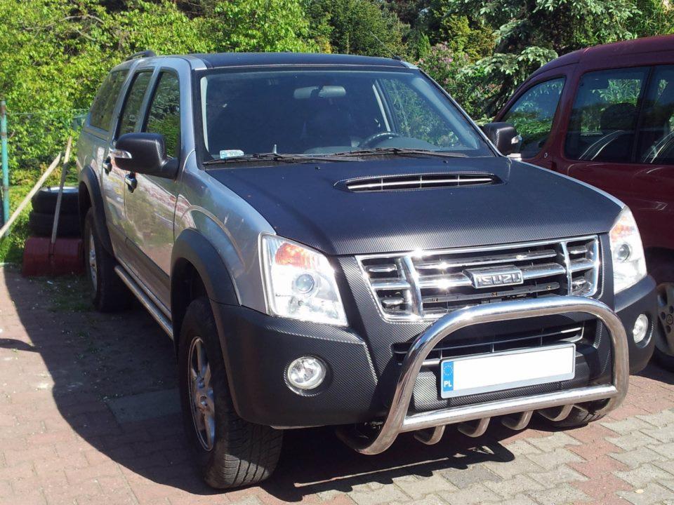 Srebrny samochód Isuzu z czarną folią imitującą karbon na masce i elementach karoserii, widoczny orurowany przedni zderzak.