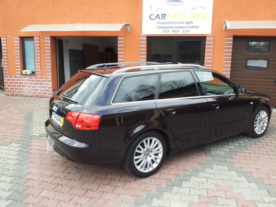 Ciemny samochód kombi Audi A4 z przyciemnionymi szybami, zaparkowany przed budynkiem warsztatu z szyldem reklamującym przyciemnianie szyb i oklejanie samochodów.