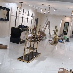 AS Firma Remontowo-Budowlana - Wnętrze sklepu odzieżowego w trakcie montażu wyposażenia: złote regały ekspozycyjne, wieszaki na ubrania, lustra w czarnych ramach, biała posadzka z połyskiem, częściowo rozpakowane kartony...