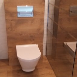 AS Firma Remontowo-Budowlana - Nowoczesna toaleta podwieszana z panelem drewnopodobnym i chromowanym przyciskiem spłukującym, kabina prysznicowa ze szklaną ścianką w tle, podłoga z paneli imitujących drewno.