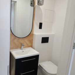 AS Firma Remontowo-Budowlana - Nowoczesna łazienka po remoncie: czarna szafka pod umywalką z owalnym lustrem i drewnianą mozaiką w tle, wisząca toaleta, minimalistyczna lampa sufitowa.
