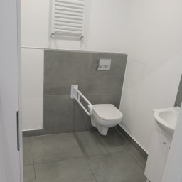 AS Firma Remontowo-Budowlana - Nowoczesna, jasna łazienka z szarymi akcentami, wyposażona w toaletę z uchwytem dla osób z niepełnosprawnościami, białą umywalkę z szafką i grzejnik drabinkowy na ścianie.