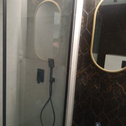 AS Firma Remontowo-Budowlana - Nowoczesna kabina prysznicowa z deszczownicą i czarną armaturą na tle heksagonalnych płytek w odcieniach brązu i złota, uzupełniona owalnym lustrem z ramą w kolorze złota.