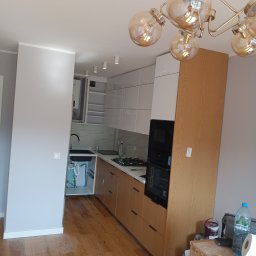 AS Firma Remontowo-Budowlana - Nowoczesna kuchnia z drewnianymi i białymi szafkami, czarnym zlewem i marmurowym backsplashiem; widoczne elementy instalacji pod zlewem, oświetlenie sufitowe i bałagan na podłodze obok szafek.