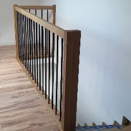 Drewniana balustrada schodowa z czarnymi tralkami, widok z góry na schody zabezpieczone kartonem i taśmą malarską, podłoga wyłożona panelami imitującymi drewno.