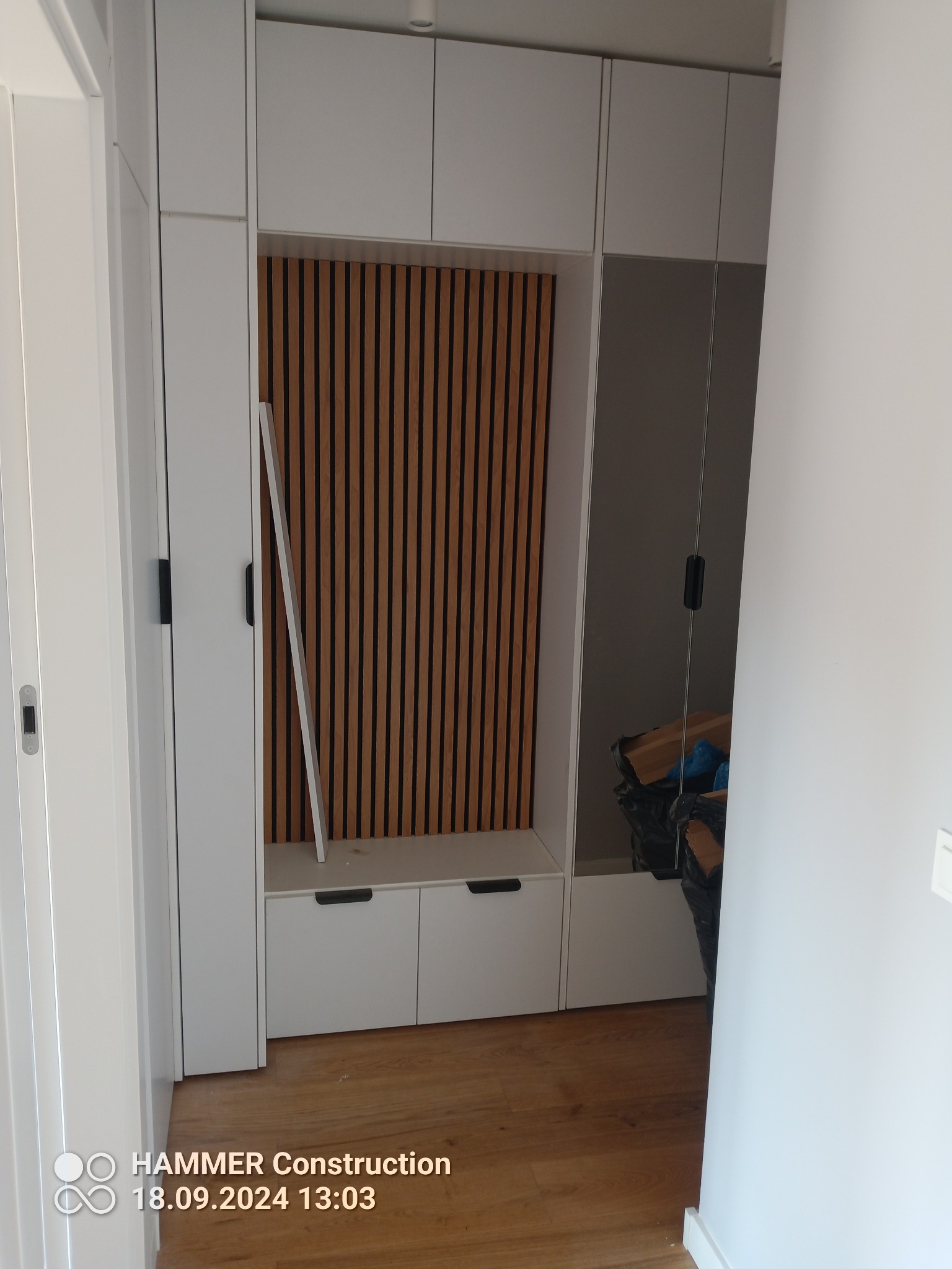 Wnęka w przedpokoju z zabudową z białych szafek i drewnianymi lamelami na ścianie, widoczny fragment lustra i podłogi z paneli.