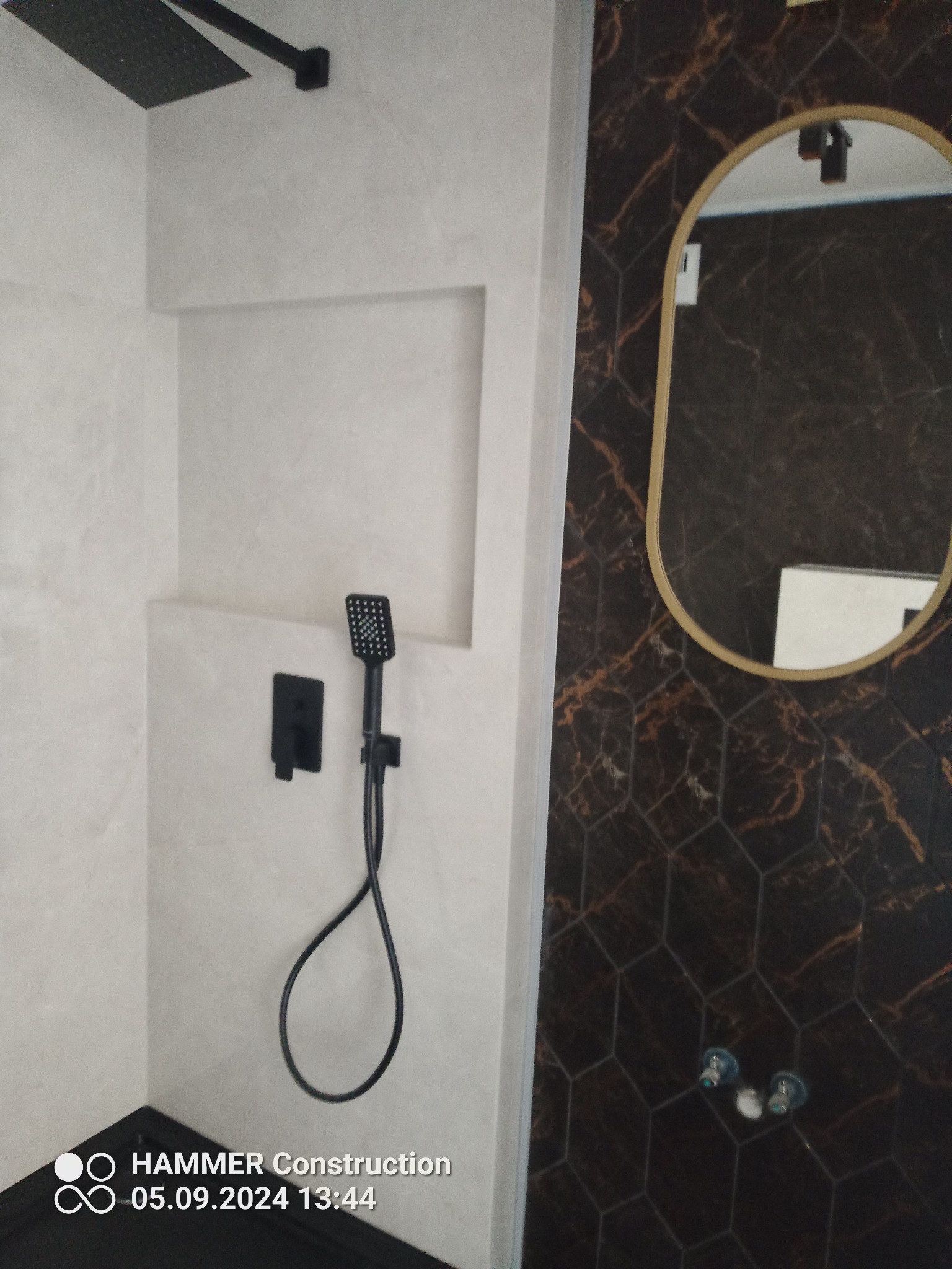 Nowoczesna łazienka z czarną armaturą: deszczownica, bateria podtynkowa i słuchawka prysznicowa na tle jasnych, imitujących beton ścian oraz ciemnych, heksagonalnych płytek z marmurkowym wzorem...