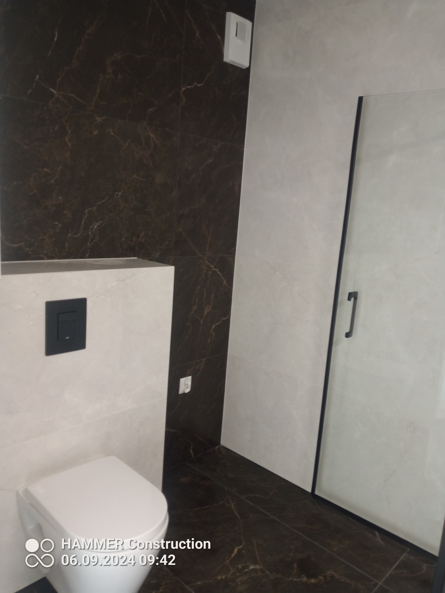 Nowoczesna łazienka z brązowymi płytkami imitującymi marmur na ścianach i podłodze, wisząca toaleta z czarnym przyciskiem spłukiwania i kabina prysznicowa z czarnymi ramami.