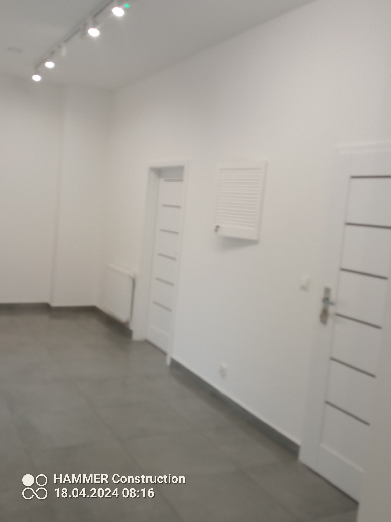 Wnętrze w trakcie prac wykończeniowych: szara podłoga, białe ściany z grzejnikiem i wentylacją, białe drzwi z poziomymi panelami, oświetlenie sufitowe.