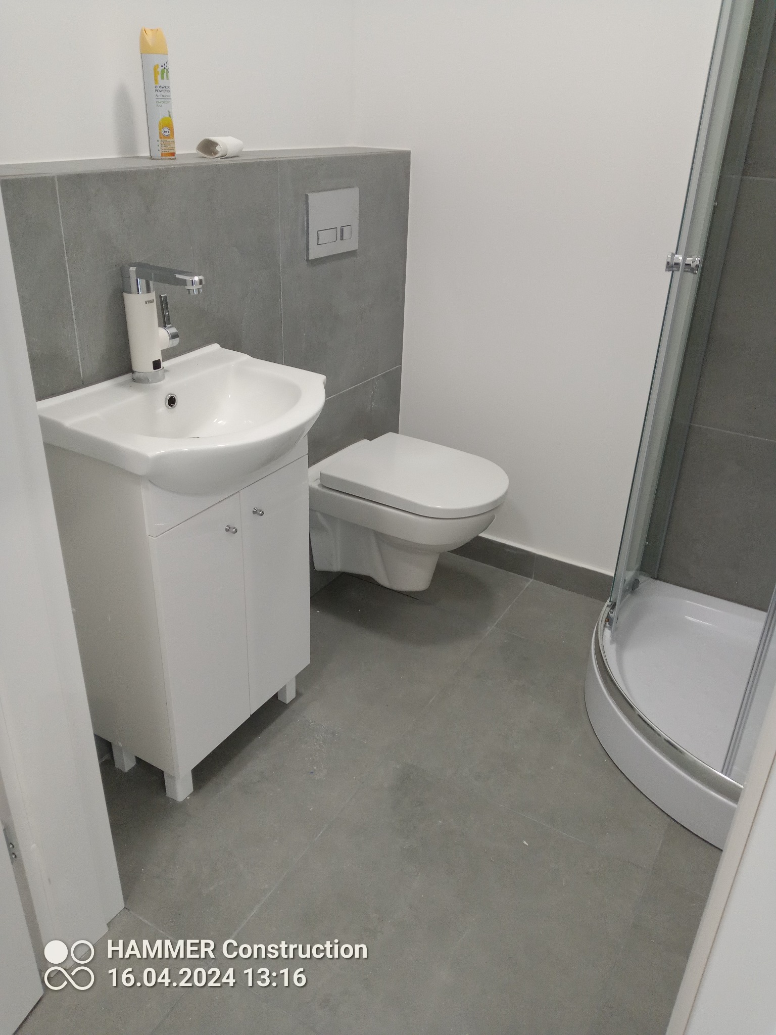 Wykończona łazienka w odcieniach szarości i bieli: umywalka z szafką, toaleta i kabina prysznicowa z zaokrąglonym frontem, widoczny dozownik na ścianie.