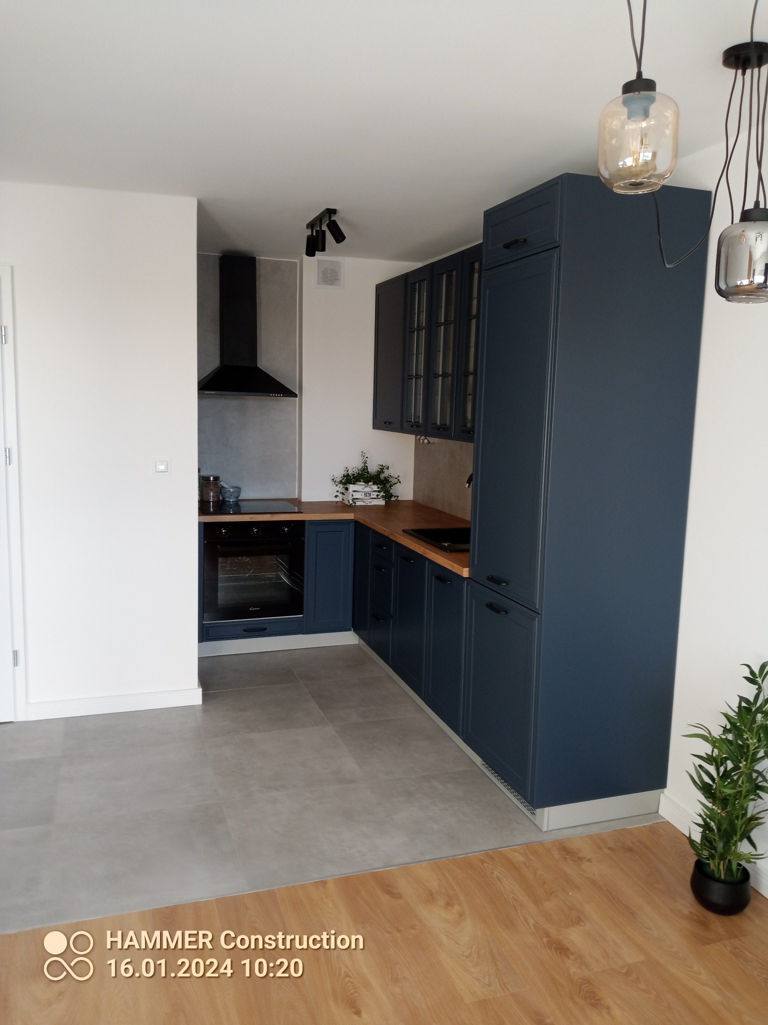 Nowoczesna kuchnia w odcieniach granatu i drewna, z wiszącymi lampami o różnorodnych kształtach, widok na płytę grzewczą i okap, podłoga wykończona płytkami imitującymi beton przechodzącymi w panele.