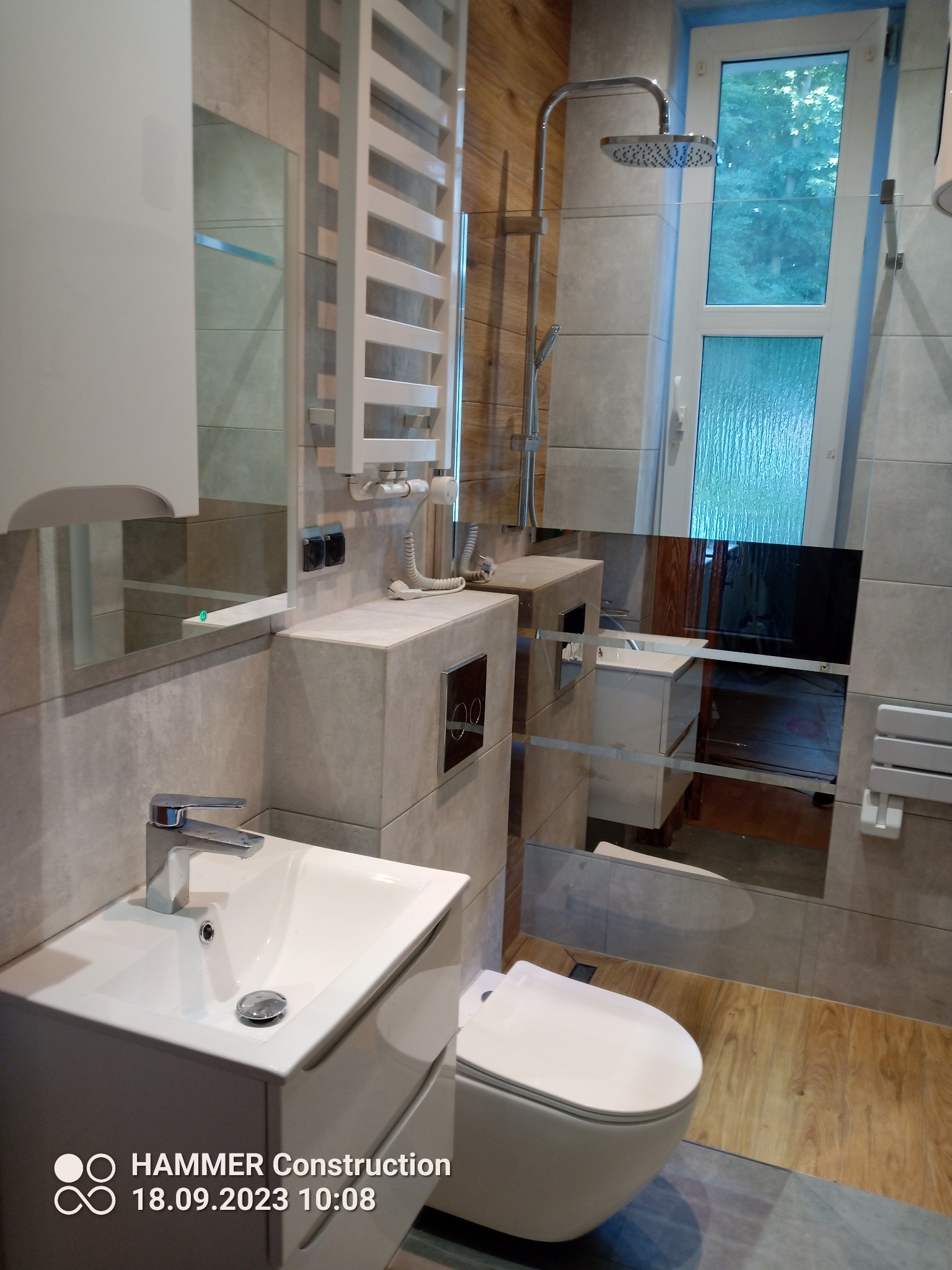 Nowoczesna łazienka z szarą glazurą, drewnopodobną ścianą i podłogą, białą armaturą, grzejnikiem drabinkowym i kabiną prysznicową typu walk-in.