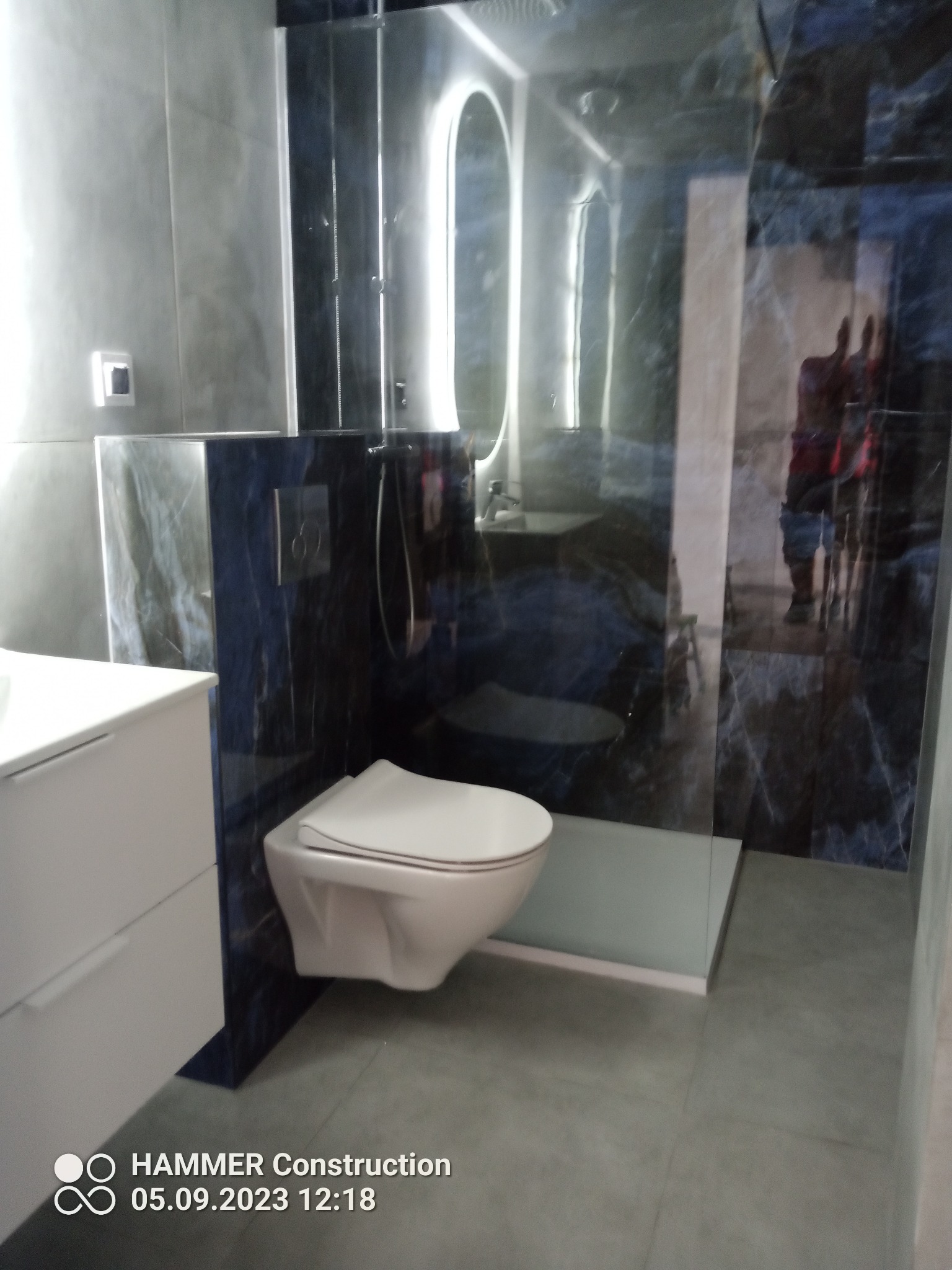 Nowoczesna łazienka z podwieszaną toaletą, kabiną prysznicową typu walk-in i umywalką na szafce, wykończona płytkami imitującymi marmur w odcieniach szarości i granatu.