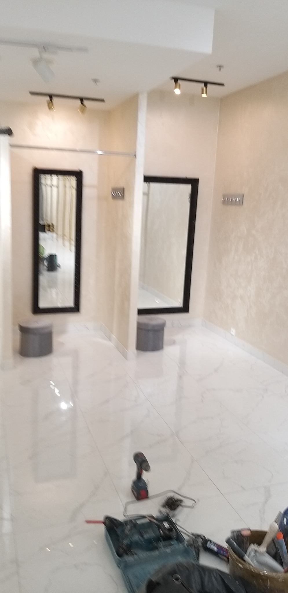 Wykończone wnętrze garderoby z lustrami w czarnych ramach, szarymi pufami i białymi marmurowymi płytkami podłogowymi; narzędzia i sprzęt glazurniczy widoczne na pierwszym planie sugerują prace...